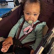Brooklynn is registered to the contest to win money with this photo: toddler, child, stroller, pacifier, purple_bow, curly_hair, blue_eyes, toy, seatbelt, clothing, long_sleeve, black_pants, baby, portrait, cute, indoor, person, sitting, infant, accessory