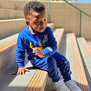 Elias is registered to the contest to win money with this photo: toddler, child, blue_clothing, sitting, stairs, outdoor, sunlight, concrete, curly_hair, white_shoes, playful, casual_wear, daylight, expression, young, person, hand, urban, background, plant