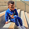 toddler, child, blue_clothing, sitting, stairs, outdoor, sunlight, concrete, curly_hair, white_shoes, playful, casual_wear, daylight, expression, young, person, hand, urban, background, plant