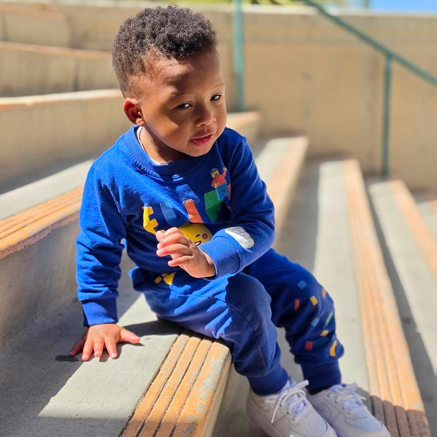 Elias is registered to the contest to win money with this photo: toddler, child, blue_clothing, sitting, stairs, outdoor, sunlight, concrete, curly_hair, white_shoes, playful, casual_wear, daylight, expression, young, person, hand, urban, background, plant