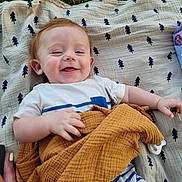 Sonny is registered to the contest to win money with this photo: baby, smiling, blanket, grass, outdoor, child, happy, clothing, face, person, pattern, blue_stripes, white_shirt, mustard_cloth, pacifier, hand, skin, head, ear, lying_down