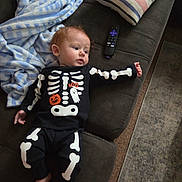 Sonny is registered to the contest to win money with this photo: baby, black_clothing, blanket, carpet, child, couch, curious, footwear_off, ghost, halloween, home, indoor, infant, pillow, pumpkin, relaxed, remote_control, skeleton_costume, striped, white_bones