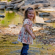 Charlea is registered to the contest to win money with this photo: child, toddler, girl, smiling, portrait, outdoor, creek, water, stream, rocks, nature, park, floral_dress, jeans, standing, reflection, sunlight, trees, pebbles, happy