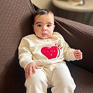 Elyana joined the competition — help win amazing prizes! baby, child, infant, sitting, clothing, heart, love, white_outfit, brown_chair, indoor, cute, portrait, soft_focus, cozy, young, person, expression, furniture, blurred_background, comfortable