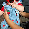 Elaine is registered to the contest to win money with this photo: baby, child, toddler, portrait, sunglasses, heart_shaped_glasses, red_bow, denim, overalls, strawberry, fashion, smiling, indoor, seated, adult_arms, support, dresser, carpet, sock, colorful_clothing