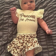 Raniah is registered to the contest to win money with this photo: baby, infant, child, headband, leopard_print, clothing, shorts, blanket, bed, lying_down, cute, expression, face, skin, person, small_hands, small_feet, fashion, text_on_clothing, indoor