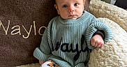 Waylon is registered to the contest to win money with this photo: baby, infant, child, sweater, pants, patterned_clothing, chair, blanket, cozy, indoor, cute, seated, portrait, blue_sweater, plush_fabric, curious_expression, small_hands, footwear, soft_lighting, home