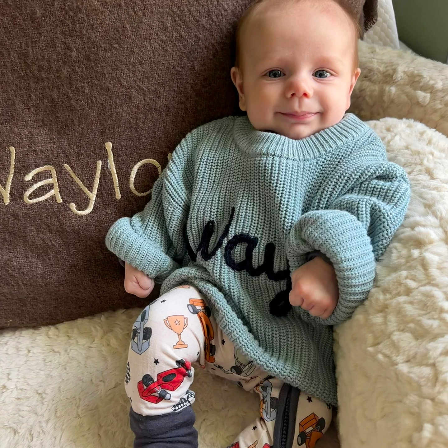 Waylon is registered to the contest to win money with this photo: baby, child, smiling, sweater, pants, patterned_clothing, chair, pillow, embroidered_text, cozy, indoor, cute, footwear, barefoot, person, portrait, seated, knitwear, happy, infant