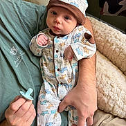 Waylon is registered to the contest to win money with this photo: baby, infant, onesie, hat, pacifier, adult_hand, green_shirt, bed, blanket, couch, animal_print, cute, portrait, indoor, soft_lighting, cozy, child, newborn, clothing, person