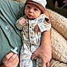 baby, infant, onesie, hat, pacifier, adult_hand, green_shirt, bed, blanket, couch, animal_print, cute, portrait, indoor, soft_lighting, cozy, child, newborn, clothing, person