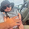 baby, infant, child, sunglasses, hat, floppy_hat, car, car_interior, steering_wheel, car_seat, sitting, portrait, face, clothing, gray_shirt, adult_hand, safety, road_trip, indoors, photography