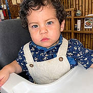Emiliano is registered to the contest to win money with this photo: child, toddler, portrait, high_chair, sitting, curly_hair, blue_eyes, floral_shirt, overalls, bib, restaurant, indoor, bamboo, wooden_wall, shelves, food_poster, camera, serious_expression, table, baby