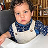 child, toddler, portrait, high_chair, sitting, curly_hair, blue_eyes, floral_shirt, overalls, bib, restaurant, indoor, bamboo, wooden_wall, shelves, food_poster, camera, serious_expression, table, baby