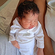 Yuhanna participe au concours pour gagner de l'argent avec cette photo : baby, newborn, infant, sleeping, lap, onesie, white_clothing, curly_hair, person, adult_arm, bracelet, hand, face, portrait, closeup, indoors, bedding, pillow, skin_tone, cozy