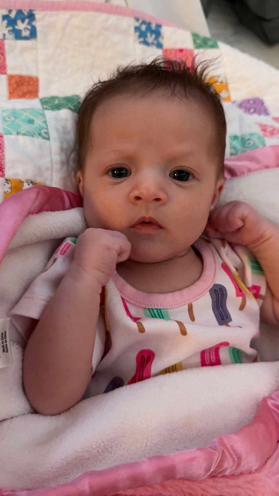 Charleigh is registered to the contest to win money with this photo: baby, infant, blanket, pink, onesie, colorful, popsicle_pattern, face, fist, curly_hair, indoor, cute, child, portrait, young, newborn, cozy, expression, skin, person