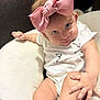 baby, infant, bow, pink_bow, onesie, white_clothing, cushion, blanket, hand, leg, face, eyes, skin, indoor, person, cute, child, sitting, expression, soft