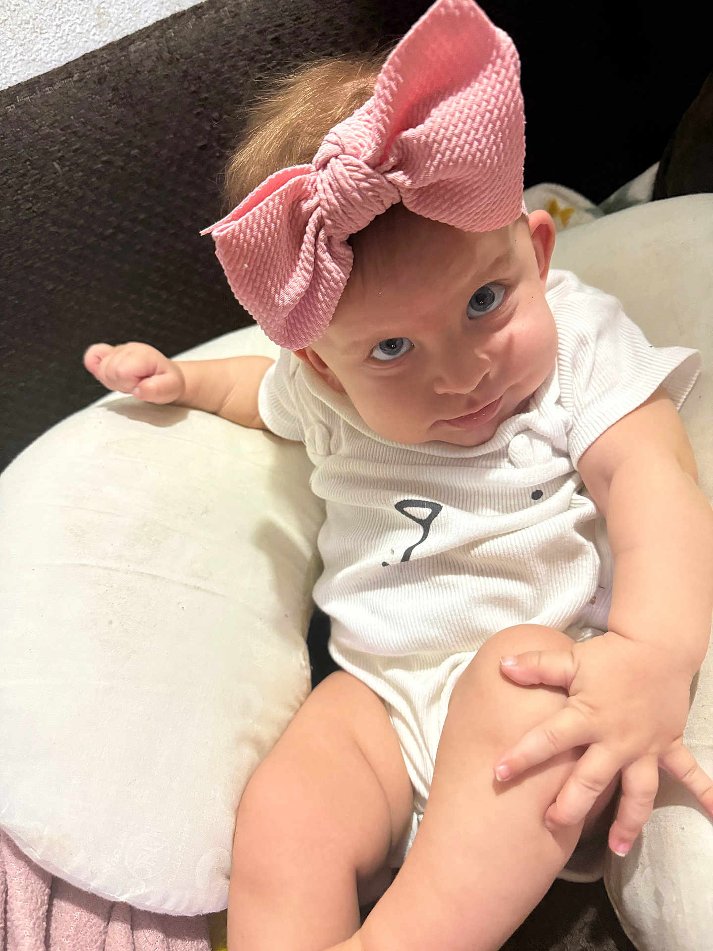 Nymeria is registered to the contest to win money with this photo: baby, infant, bow, pink_bow, onesie, white_clothing, cushion, blanket, hand, leg, face, eyes, skin, indoor, person, cute, child, sitting, expression, soft