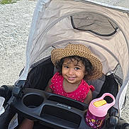 Esmè is registered to the contest to win money with this photo: toddler, child, stroller, hat, smile, curly_hair, pink_dress, sippy_cup, outdoor, park, lake, grass, tree, sunny, path, water, nature, happy, baby_shoes, shade