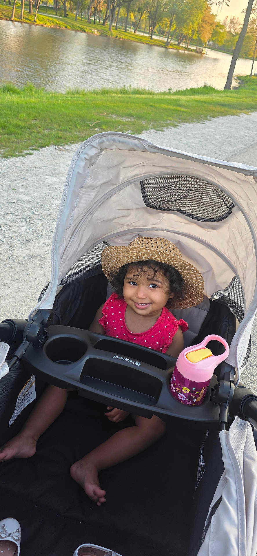 Esmè is registered to the contest to win money with this photo: toddler, child, stroller, hat, smile, curly_hair, pink_dress, sippy_cup, outdoor, park, lake, grass, tree, sunny, path, water, nature, happy, baby_shoes, shade