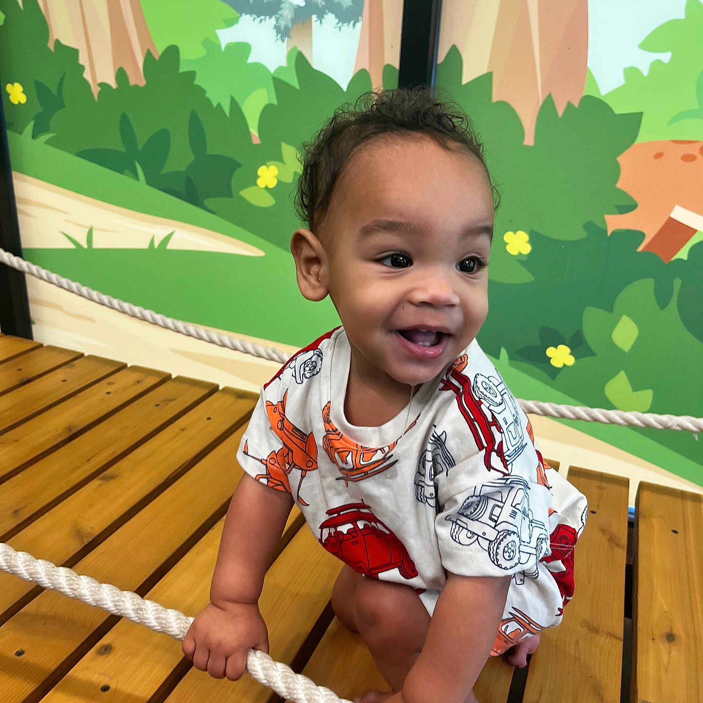 Asy joined the competition — help win amazing prizes! barefoot, bright_colors, candid, cartoon_background, child, colorful_shirt, curly_hair, cute, fun, greenery, hand_grip, happy, indoor_play_area, playful, rope, smiling, toddler, vehicles_print, wooden_platform, young_child