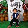 baby, backdrop, balloons, barefoot, birthday, brown, celebration, chair, child, decorations, first_birthday, football, football_theme, green, indoor, portrait, sitting, smiling, toddler, white
