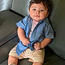 baby, child, boy, couch, leather, blue_shirt, striped_shirt, beige_shorts, white_socks, sitting, indoors, furniture, cute, portrait, young_child, curly_hair, comfortable, casual_clothing, home, expression
