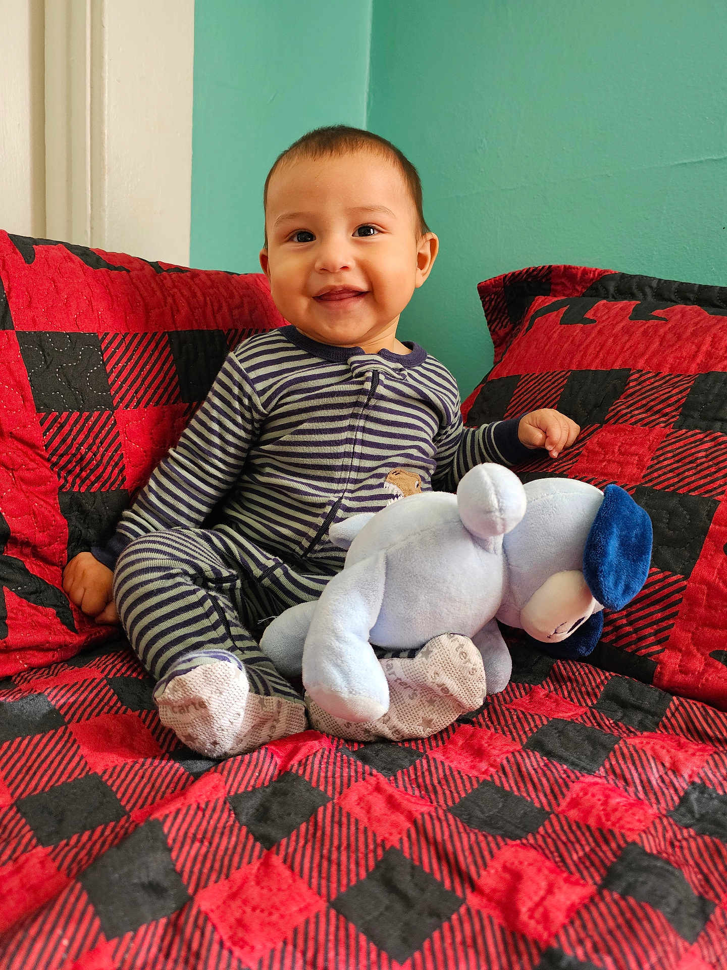 Joshua is registered to the contest to win money with this photo: baby, child, striped_pajamas, smiling, stuffed_animal, plush_toy, couch, red_and_black, checkered_pattern, sitting, indoor, teal_wall, happy, cute, infant, furniture, cozy, home, portrait, comfort