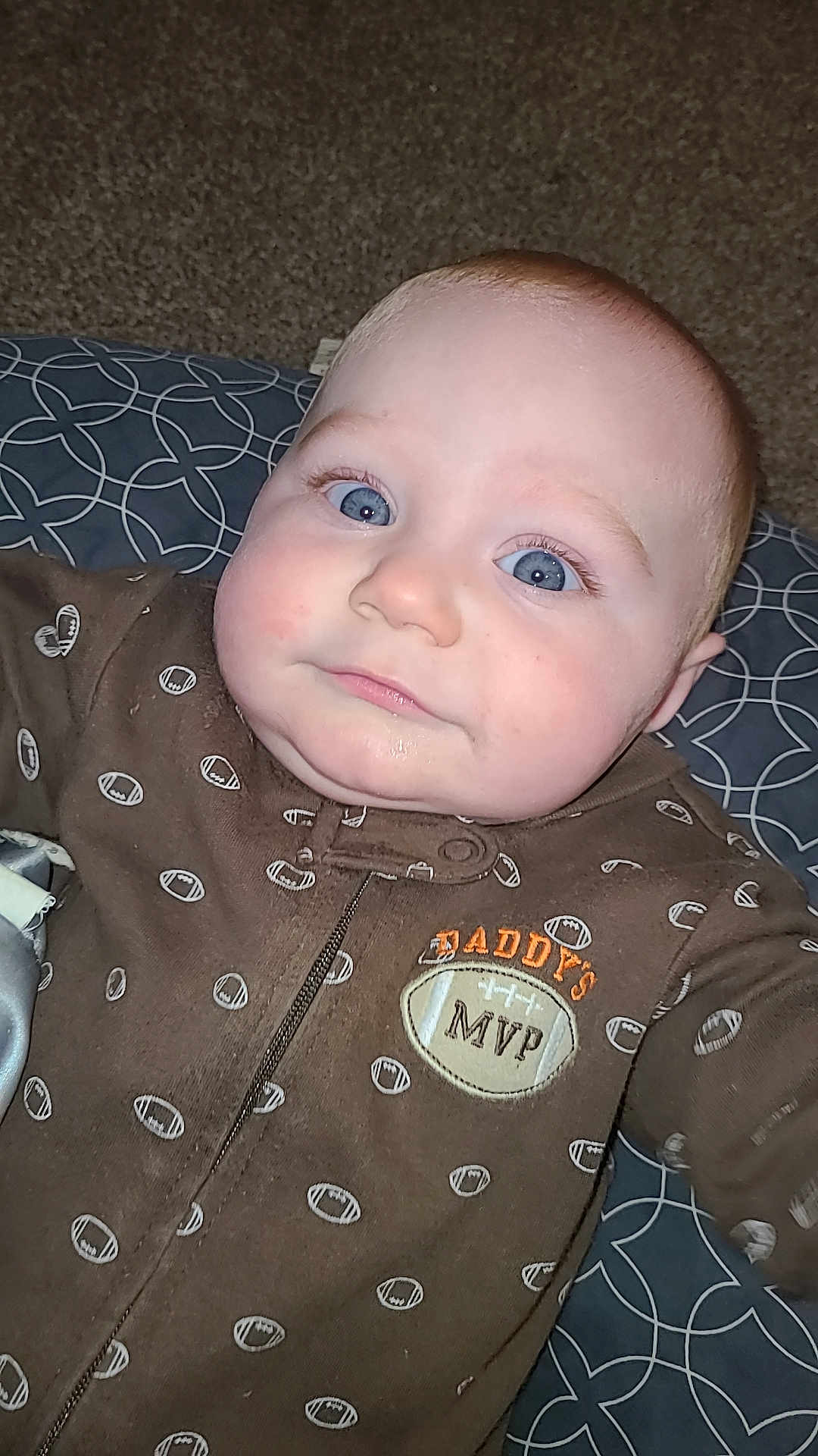 Axton is registered to the contest to win money with this photo: baby, infant, face, blue_eyes, brown_onesie, football_pattern, text_on_clothing, cushion, carpet, closeup, child, indoor, person, cute, portrait, sleepwear, young_child, smiling, head, skin