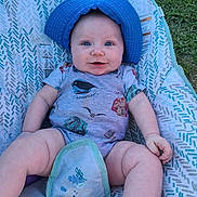Axton is registered to the contest to win money with this photo: baby, blue_hat, smiling, onesie, outdoor, grass, baby_seat, patterned_fabric, cute, chubby_legs, happy, infant, child, person, face, summer, sunlight, casual, playful, relaxed