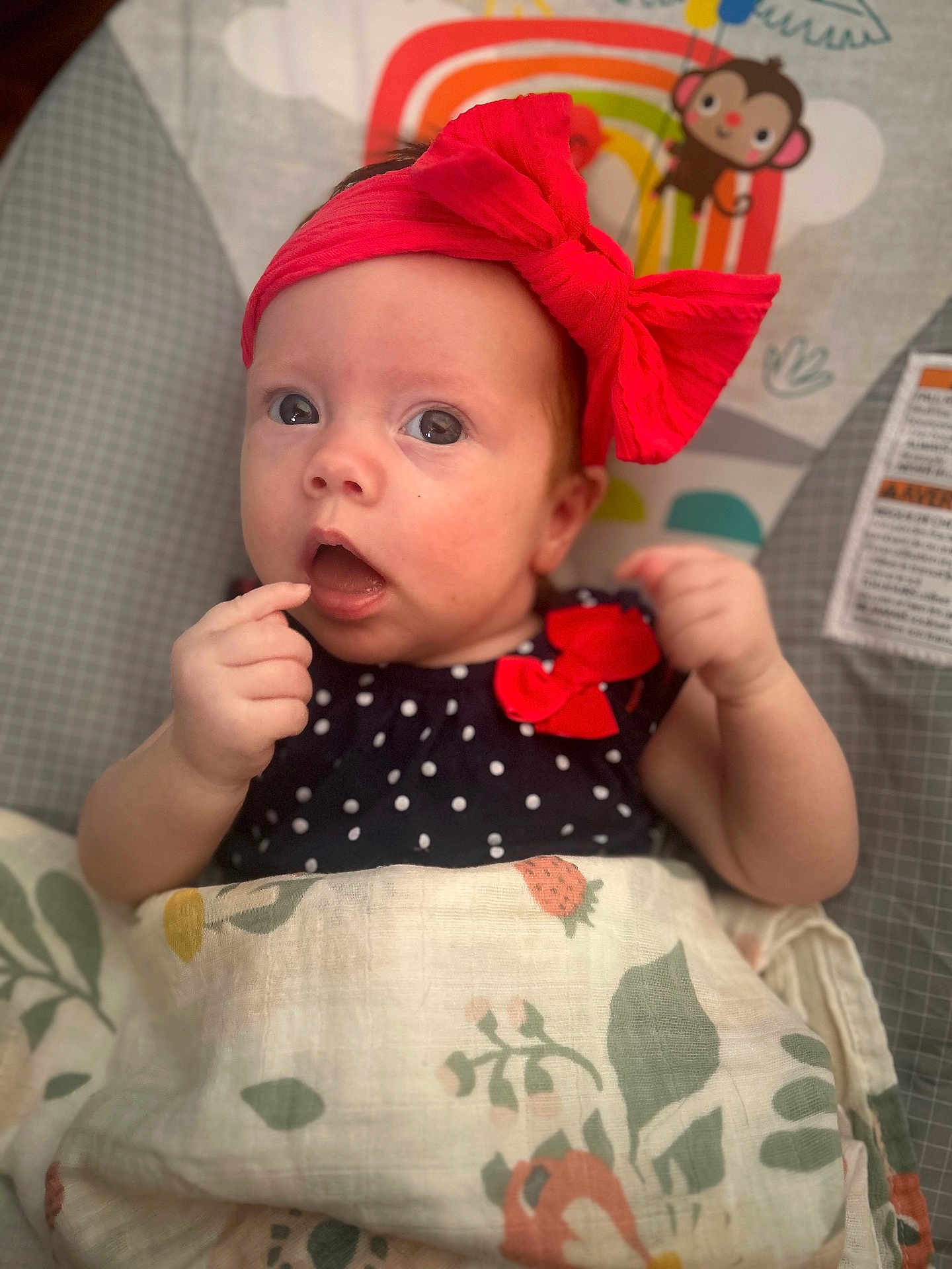 Josie is registered to the contest to win money with this photo: baby, infant, red_bow, blanket, patterned_blanket, polka_dot_clothing, monkey_illustration, baby_clothing, face, hand, indoor, cute, child, portrait, expression, newborn, headband, sitting, curious, wrapped