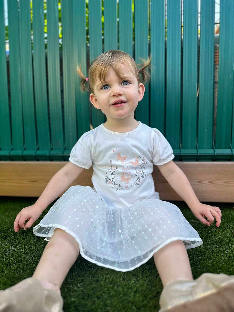 Esmee a rejoint le concours — aidez-le/la à gagner de superbes lots ! child, toddler, girl, pigtails, white_shirt, white_skirt, grass, outdoor, fence, sitting, happy, cute, young_child, portrait, smiling, daylight, casual_clothing, playful, nature, closeup