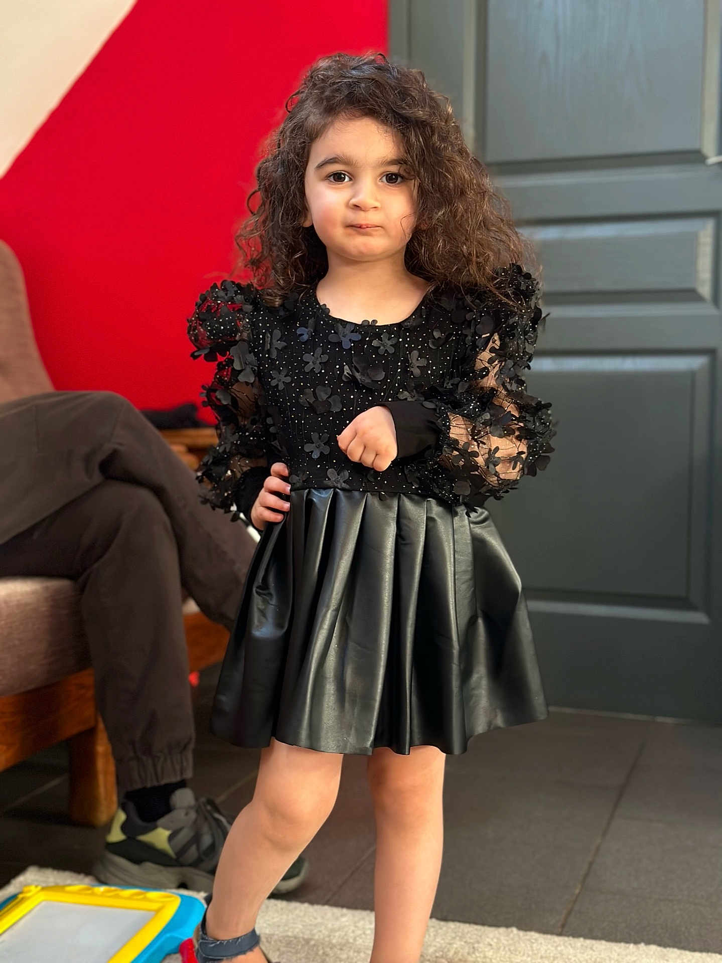 Sara a rejoint le concours — aidez-le/la à gagner de superbes lots ! child, girl, curly_hair, black_dress, indoor, red_wall, grey_door, person, floor, carpet, shoe, casual_clothing, posing, portrait, smile, hand_on_hip, furniture, sitting, toy, light
