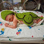Aza is registered to the contest to win money with this photo: newborn, baby, incubator, hospital, medical_tubes, crocheted_outfit, green_hat, green_shell, sleeping, premature, infant, bed, blanket, medical_equipment, turtle_costume, cute, child, healthcare, person, nursery