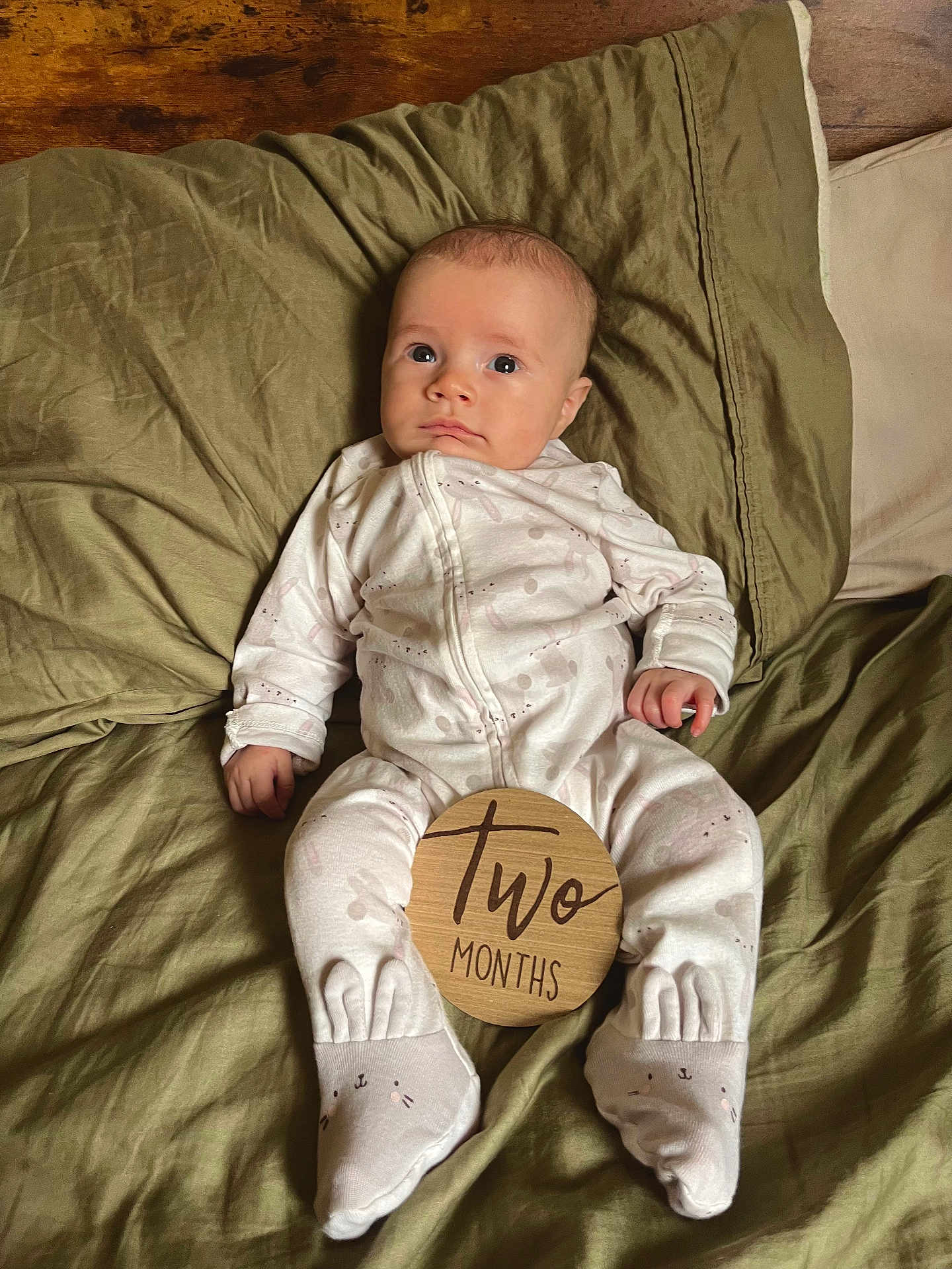 Luka is registered to the contest to win money with this photo: baby, infant, two_months, milestone, wooden_sign, onesie, blanket, pillow, bed, socks, portrait, indoors, cute, wide_eyes, hands, feet, awake, wood_floor, cozy, soft_lighting
