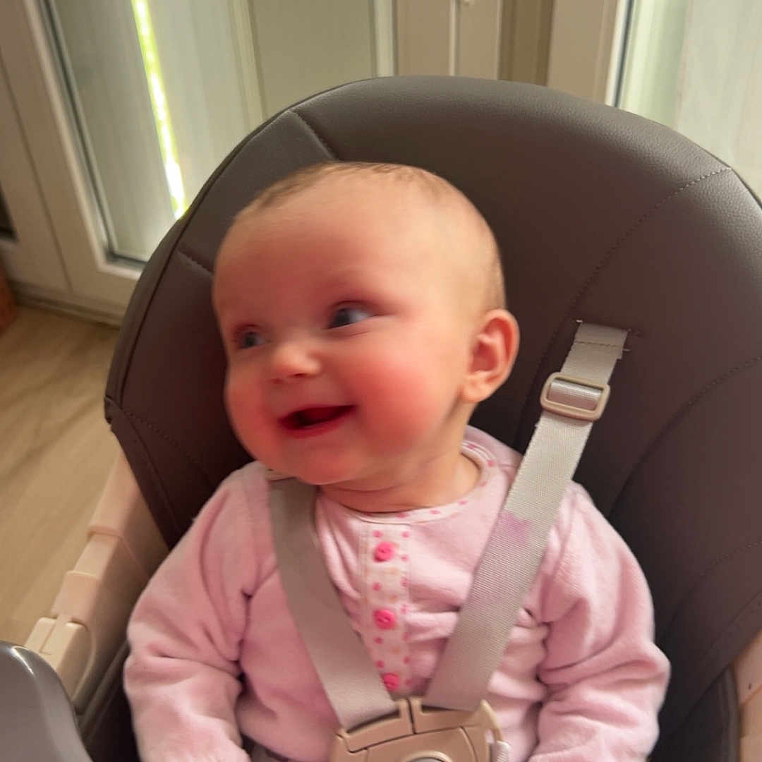Leyah a rejoint le concours — aidez-le/la à gagner de superbes lots ! baby, baby_girl, child, cute, daylight, face, furniture, hands, happy, head, high_chair, indoor, infant, person, pink_clothing, seatbelt, sitting, smiling, window, wood_floor