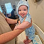 baby, infant, child, person, hands, blue_clothing, hooded_outfit, tongue_out, changing_table, baby_wipes, indoor, living_room, clock, arm, smile, cute, skin, furniture, wood_floor, adult