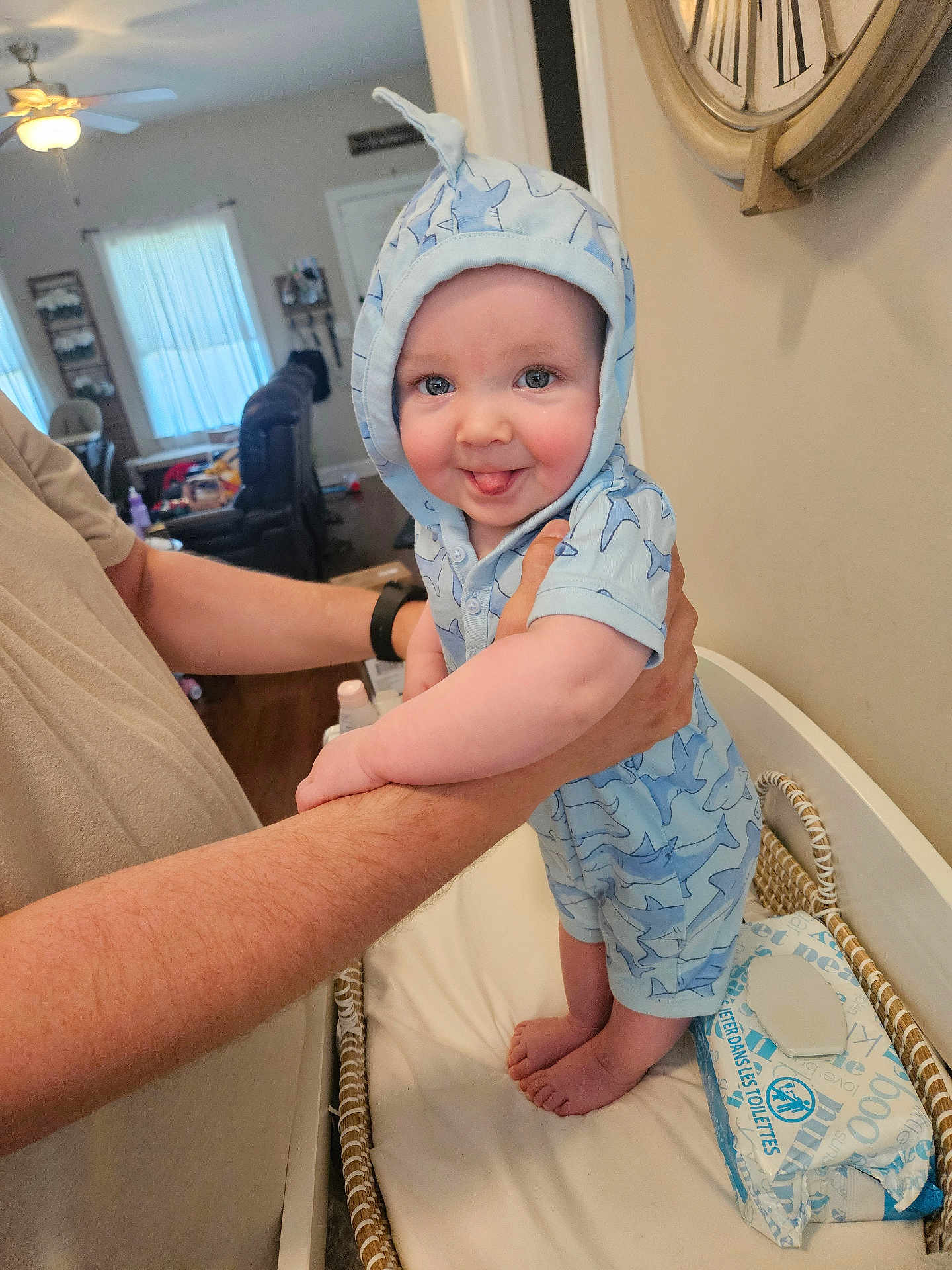 Ryder is registered to the contest to win money with this photo: baby, infant, child, person, hands, blue_clothing, hooded_outfit, tongue_out, changing_table, baby_wipes, indoor, living_room, clock, arm, smile, cute, skin, furniture, wood_floor, adult