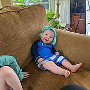 Ryder joined the competition — help win amazing prizes! baby, child, couch, living_room, smile, blue_clothing, hat, plant, lamp, window, natural_light, indoor, happy, sitting, furniture, person, legs, arm, skin, relaxing