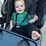 baby, child, smile, stroller, seatbelt, foot, green_shirt, cartoon_print, outdoor, happy, person, clothing, black_pants, hand, face, head, hair, toy_cart, seat, playful