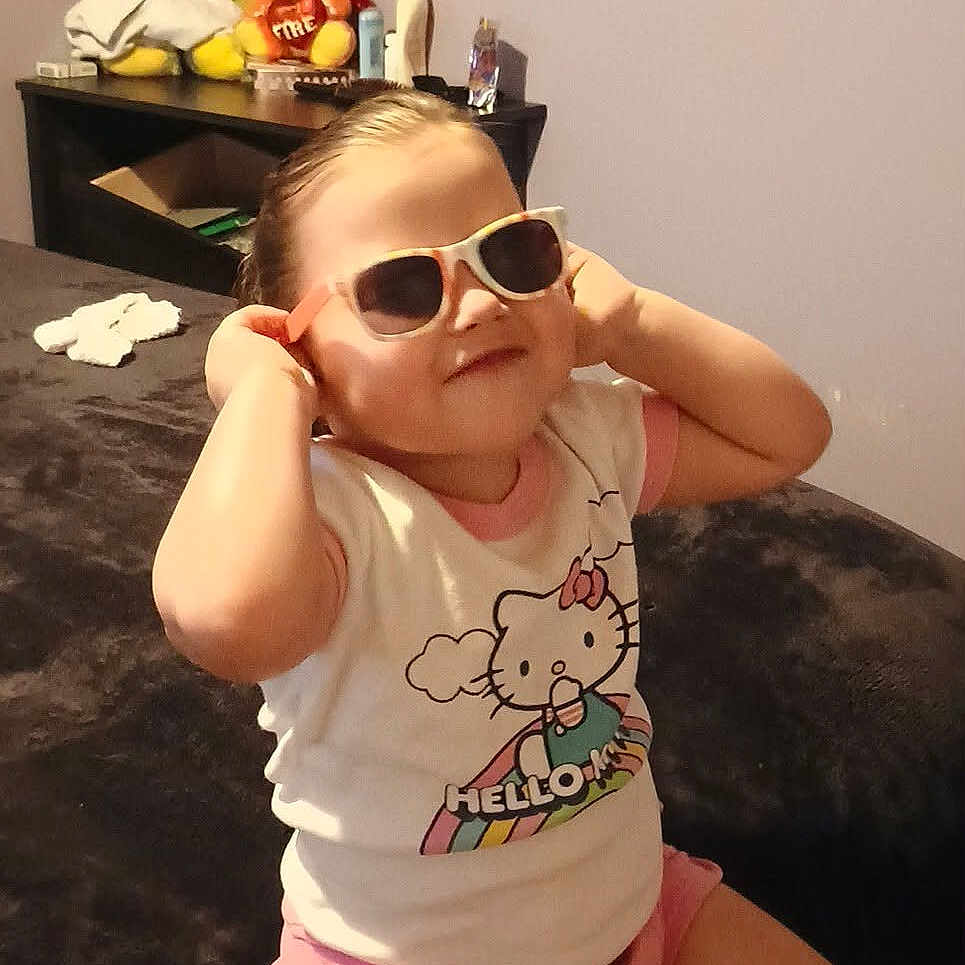 Ashley is registered to the contest to win money with this photo: child, sunglasses, hello_kitty, pink_shorts, bed, plush_toys, stuffed_animals, room, furniture, smiling, cute, indoor, casual_clothing, shorts, tshirt, person, seated, kid, playful, cozy