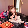 child, kid, food, fries, chicken_nuggets, ketchup, tray, bed, blanket, sitting, snack, long_hair, clothing, white_pants, black_shirt, indoor, purple_wall, bag, door, juice_pouch