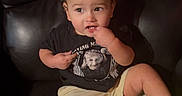Jesus is registered to the contest to win money with this photo: black_chair, casual_clothing, child, close_up, curly_hair, einstein_shirt, finger_in_mouth, furniture, indoor, leather_chair, neon_shoes, photography, playful, portrait, shorts, sitting, smiling, sneakers, t_shirt, toddler