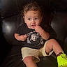 Jesus is registered to the contest to win money with this photo: toddler, child, portrait, curly_hair, smiling, sitting, leather_chair, black_chair, t_shirt, shorts, neon_shoes, sneakers, casual_clothing, indoor, furniture, close_up, playful, finger_in_mouth, einstein_shirt, photography