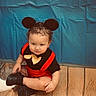 child, toddler, baby, portrait, sitting, cross_legged, costume, mouse_ears, red_clothing, black_clothing, bow_tie, curly_hair, smiling, face, indoors, blue_background, wooden_floor, shoes, fashion, photography