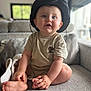 baby, child, hat, blue_eyes, beige_clothing, cactus_patch, sitting, couch, gray_couch, indoors, living_room, curious, happy, cute, portrait, infant, cozy, smiling, hands, feet