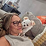 woman, glasses, dog, small_dog, white_dog, couch, blanket, pillow, indoor, casual, smile, affection, pet, shoulder, relaxation, home, cozy, blonde_hair, eyewear, portrait