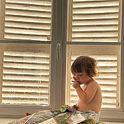 Raoul participe au concours pour gagner de l'argent avec cette photo : toddler, child, baby, book, window, shutters, pacifier, indoor, sunlight, curly_hair, sitting, reading, diaper, home, couch, window_handle, daylight, cute, curious, relaxed