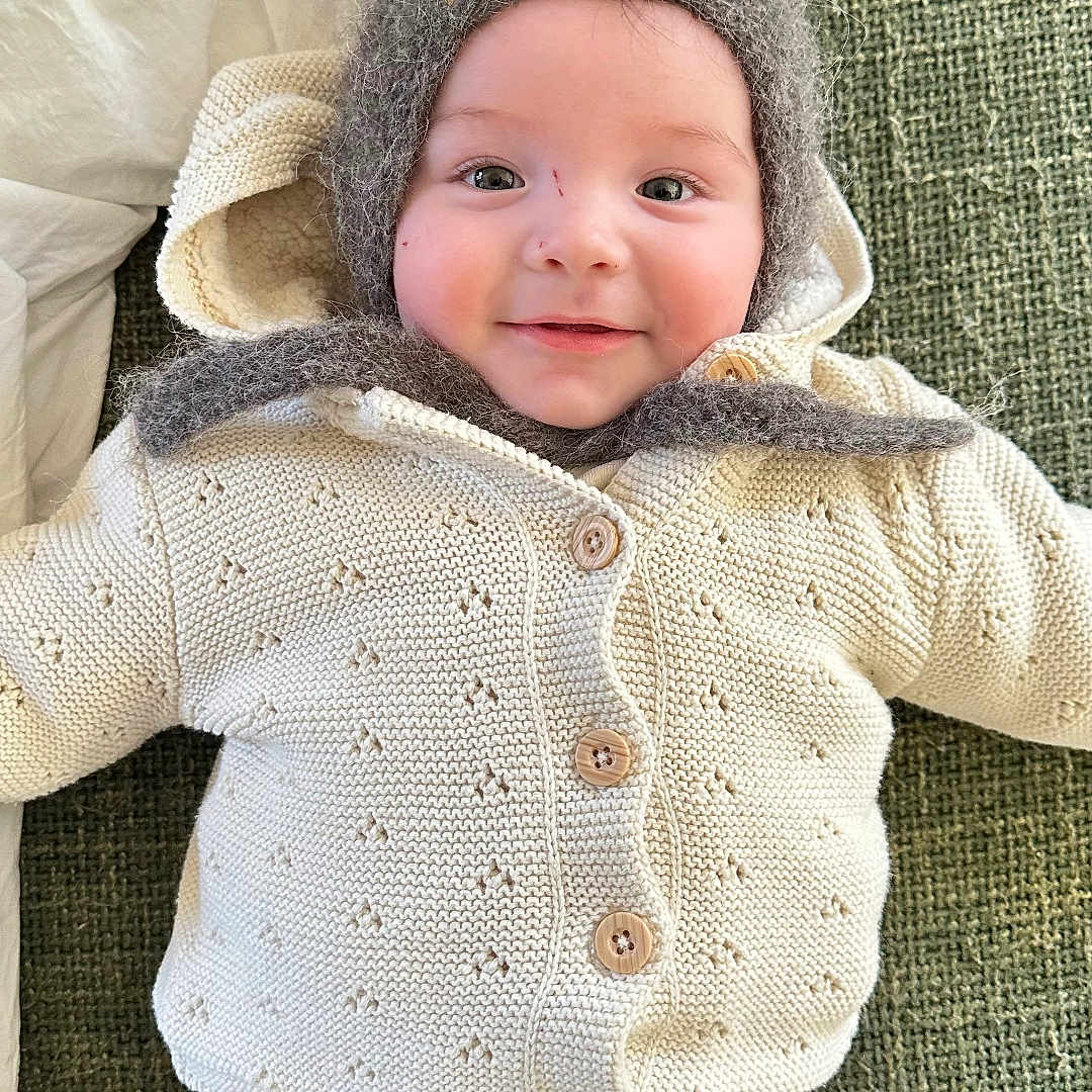 Raoul a rejoint le concours — aidez-le/la à gagner de superbes lots ! baby, buttoned_sweater, child, cozy, cute, ears, face, green_background, happy, hat, headwear, indoors, infant, knit_sweater, lying_down, pillow, portrait, smiling, soft_texture, warm_clothing