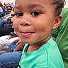child, toddler, curly_hair, green_shirt, indoor, smile, face, person, seated, people, audience, event, casual_clothing, cute, portrait, happy, young, candid, expression, human