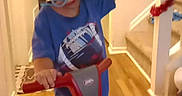 Jayden is registered to the contest to win money with this photo: blue_shirt, child, garland, happy, helmet, home, indoor, pacifier, person, playtime, railing, safety_gear, scooter, smiling, stairs, toddler, toy, white_pants, wooden_floor, young_child